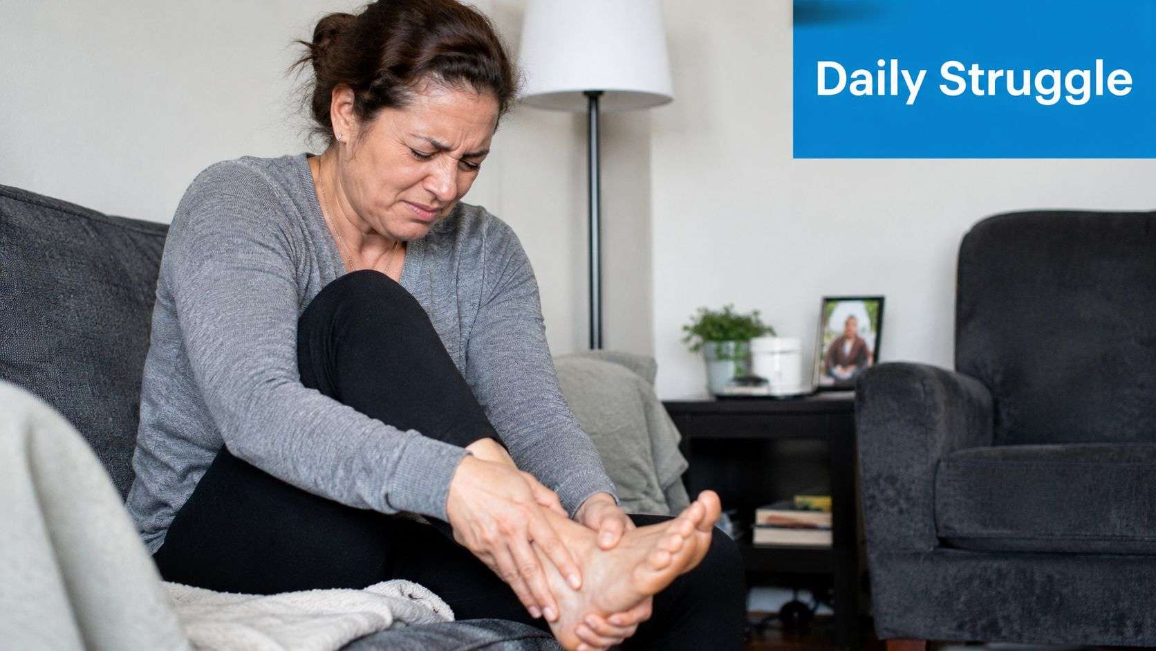 A woman sitting on a couch expressing pain while holding her foot with both hands.